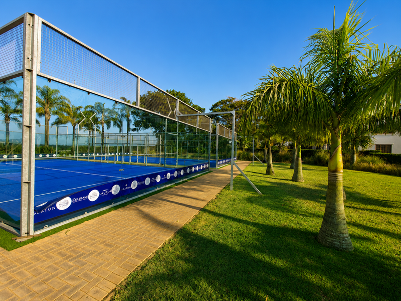 A well-maintained outdoor padel court featuring glass perimeter fencing and a paved walkway, set within a landscaped garden area.