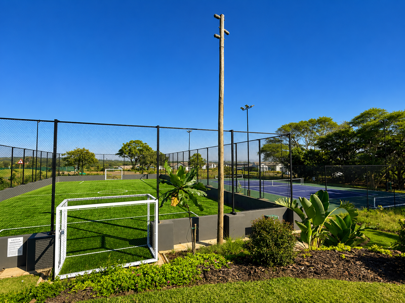 Well-maintained outdoor sports facility featuring an artificial turf soccer field and a hard-surface tennis court, enclosed by secure perimeter fencing.