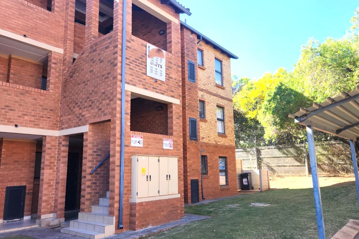 Rear view of a multi-story face brick apartment building featuring external staircases, a covered carport, and surrounding landscaped grounds.