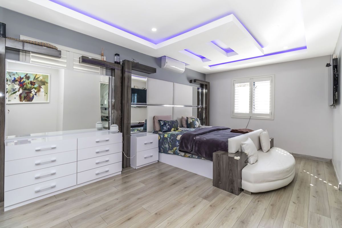 Spacious bedroom featuring a modern recessed tray ceiling with integrated LED lighting, wood-look flooring, and built-in cabinetry with mirrored accents.