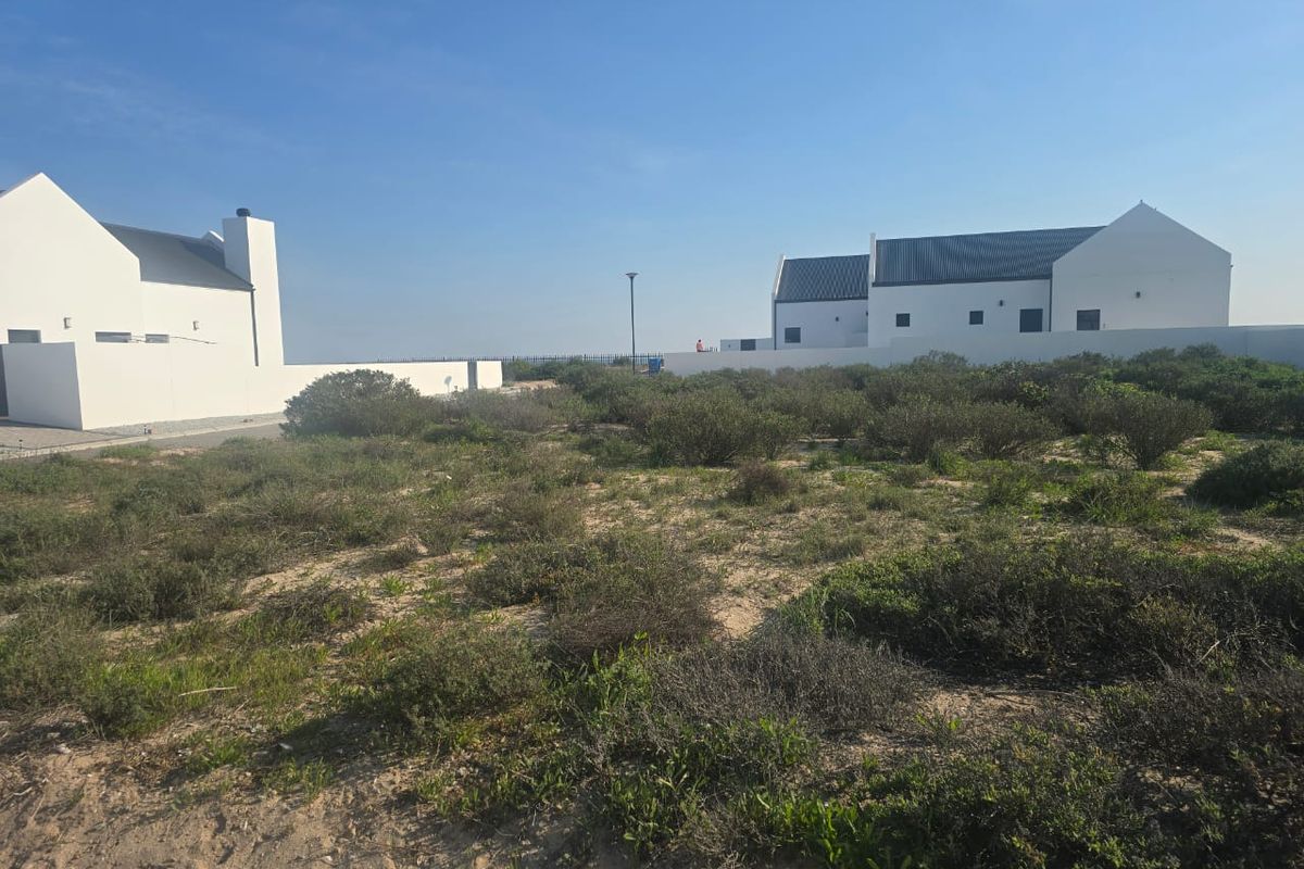 A view of a vacant residential plot featuring natural coastal vegetation with established white-walled properties in the background.