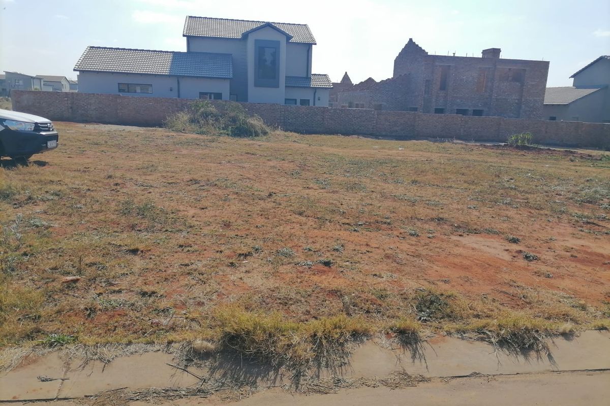 A vacant residential plot of land featuring natural ground cover, bordered by a perimeter brick wall with neighboring residential properties in the background.