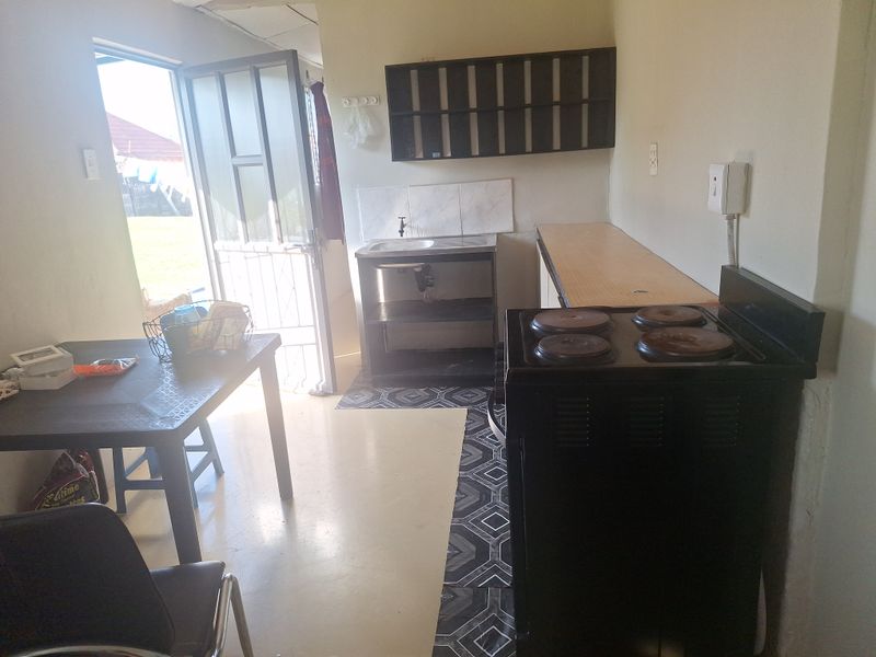 A functional kitchen area featuring a basic sink, electric stove, and wall-mounted shelving with direct access to the exterior via a security gate.
