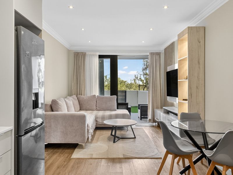 Modern open-plan living area featuring timber laminate flooring, recessed downlighting, and a built-in shelving unit, with direct access to a private balcony.