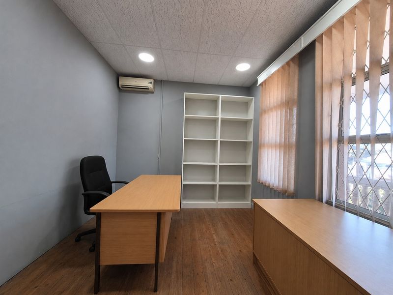 A functional office space featuring wood-look flooring, built-in shelving, vertical blinds, and a wall-mounted air conditioning unit.