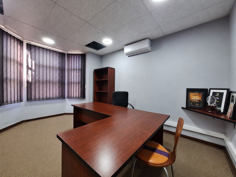 A functional office space featuring a suspended ceiling, wall-mounted air conditioning unit, and vertical window blinds.