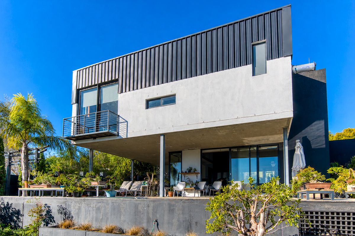 Modern two-story residence featuring a contemporary metal roof, expansive ground-floor patio, and an upper-level balcony with sliding glass door access.