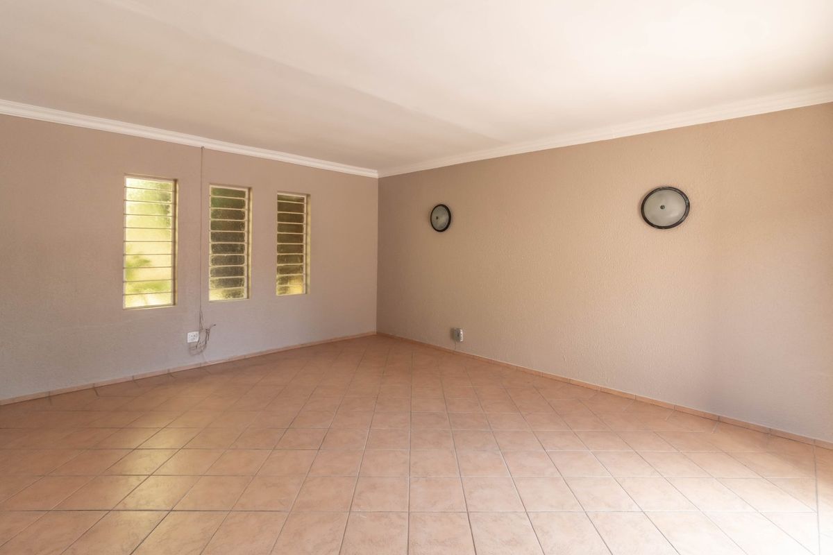 A spacious, empty living area featuring neutral wall tones, tiled flooring, and three louvered windows providing natural light.