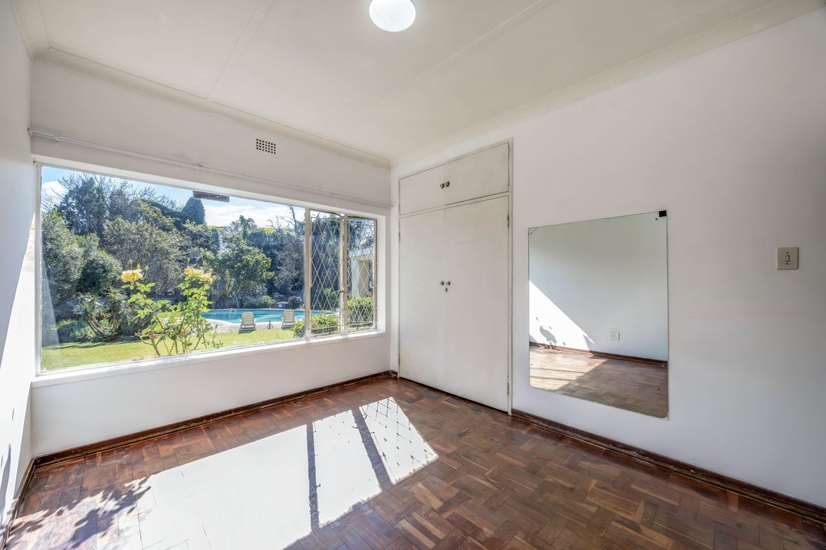 A bright, empty bedroom featuring original parquet flooring, built-in cupboards, and a large window overlooking the garden and pool area.