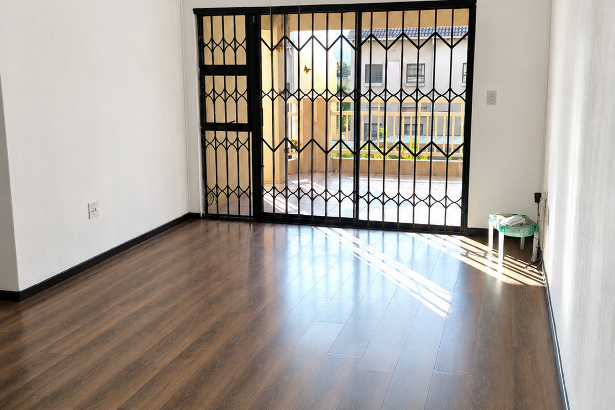 A bright, empty living room featuring wood-look laminate flooring, a ceiling fan, and a sliding glass door with a security gate leading to a balcony.