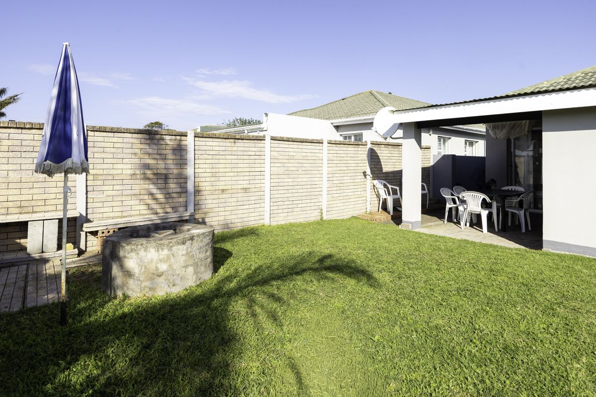 A private rear garden featuring a lawn, a built-in concrete braai pit, and a covered patio area, enclosed by a brick boundary wall.