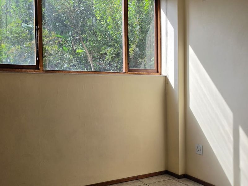 A bright, empty bedroom featuring tiled flooring and a large timber-framed window overlooking greenery.