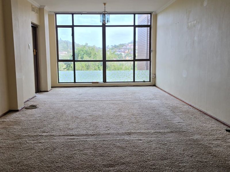 An empty, carpeted living room featuring a large window with natural light and views, requiring cosmetic updates to the walls and flooring.
