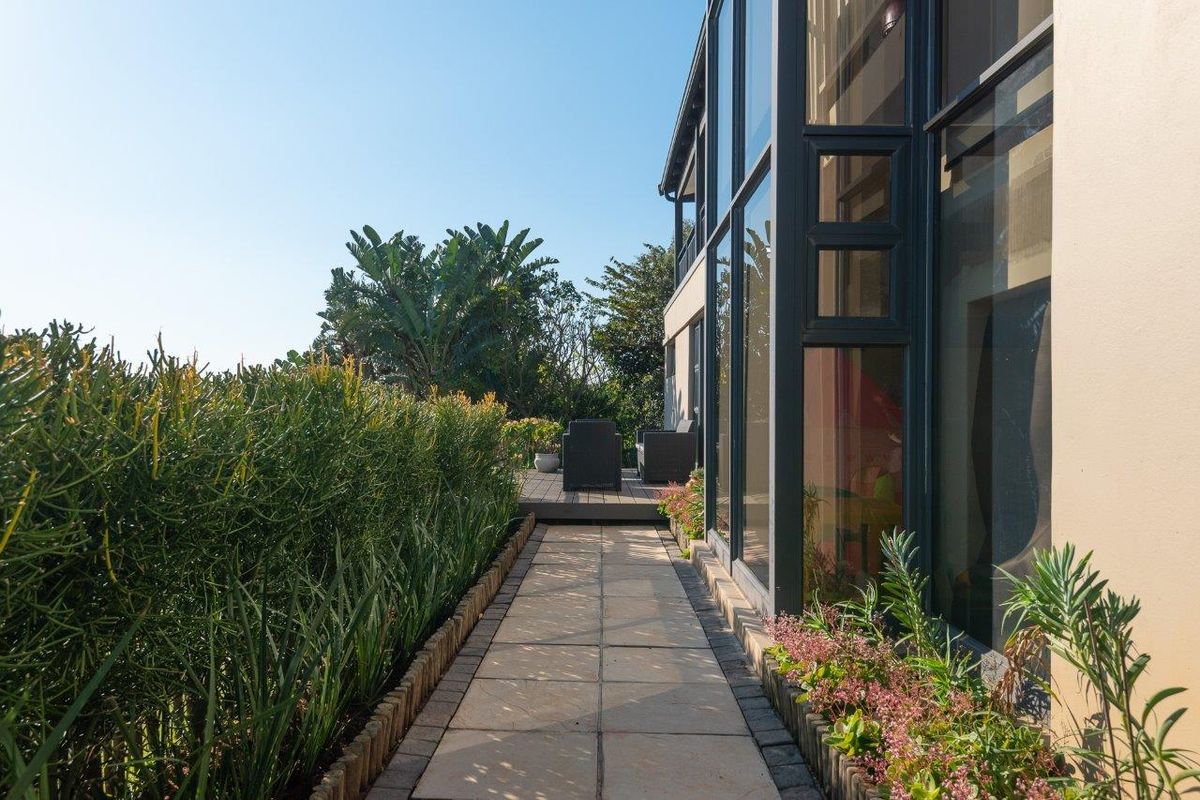 A well-maintained exterior rear pathway featuring paved stone tiles, landscaped garden borders, and large floor-to-ceiling glass windows along the building facade.