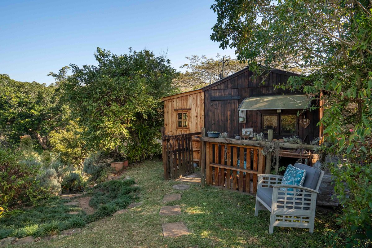 A rustic timber-clad structure featuring a small deck and awning, set within a lush, landscaped garden environment.