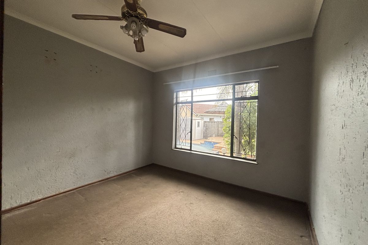 An empty bedroom featuring a ceiling fan, large window with security bars, and carpeted flooring, overlooking the rear garden area.