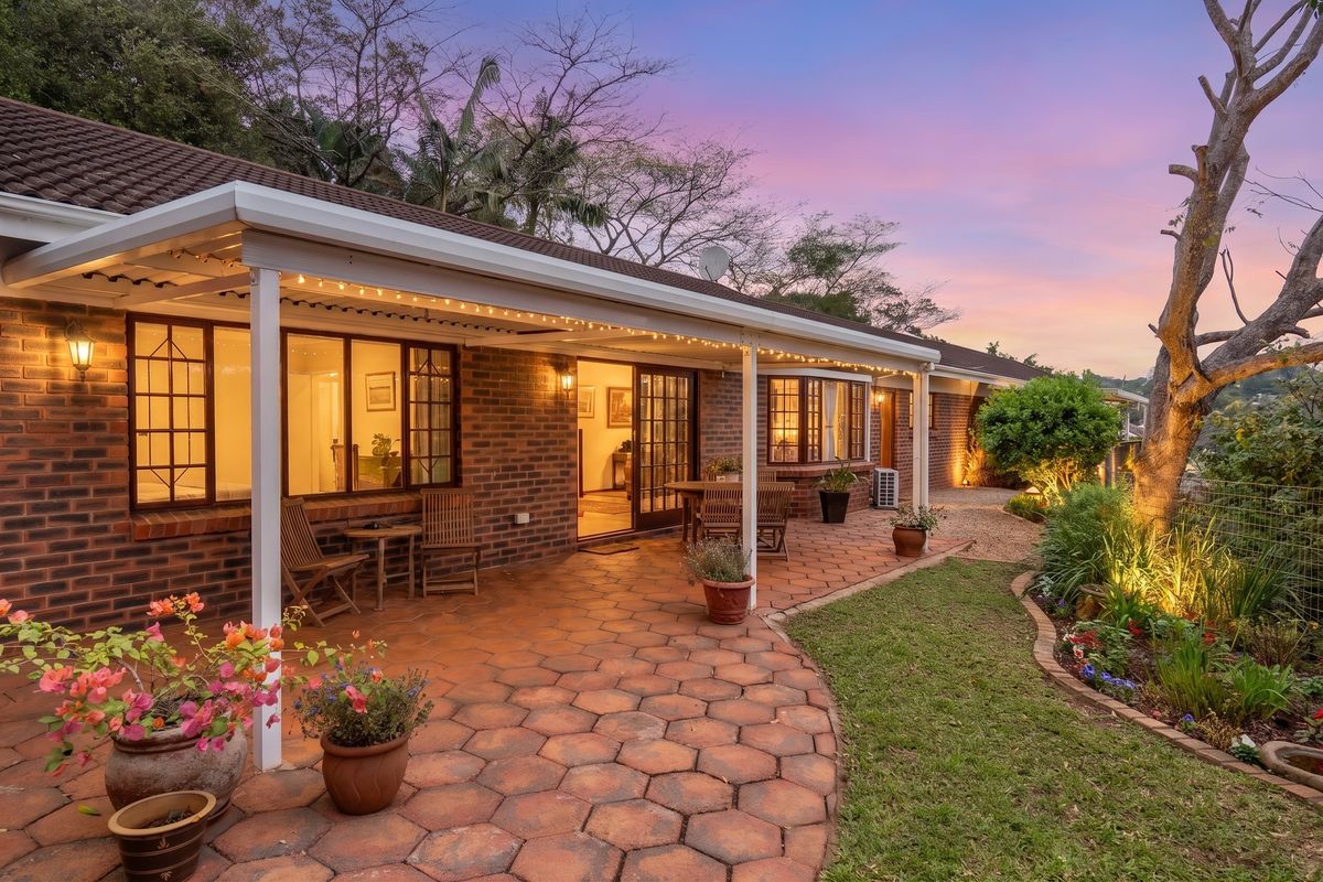A well-maintained rear exterior featuring a spacious covered patio with brick paving, landscaped garden beds, and warm evening lighting.