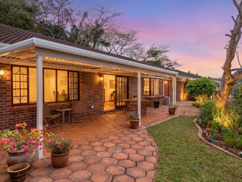 A well-maintained rear exterior featuring a spacious covered patio with brick paving, landscaped garden beds, and warm evening lighting.