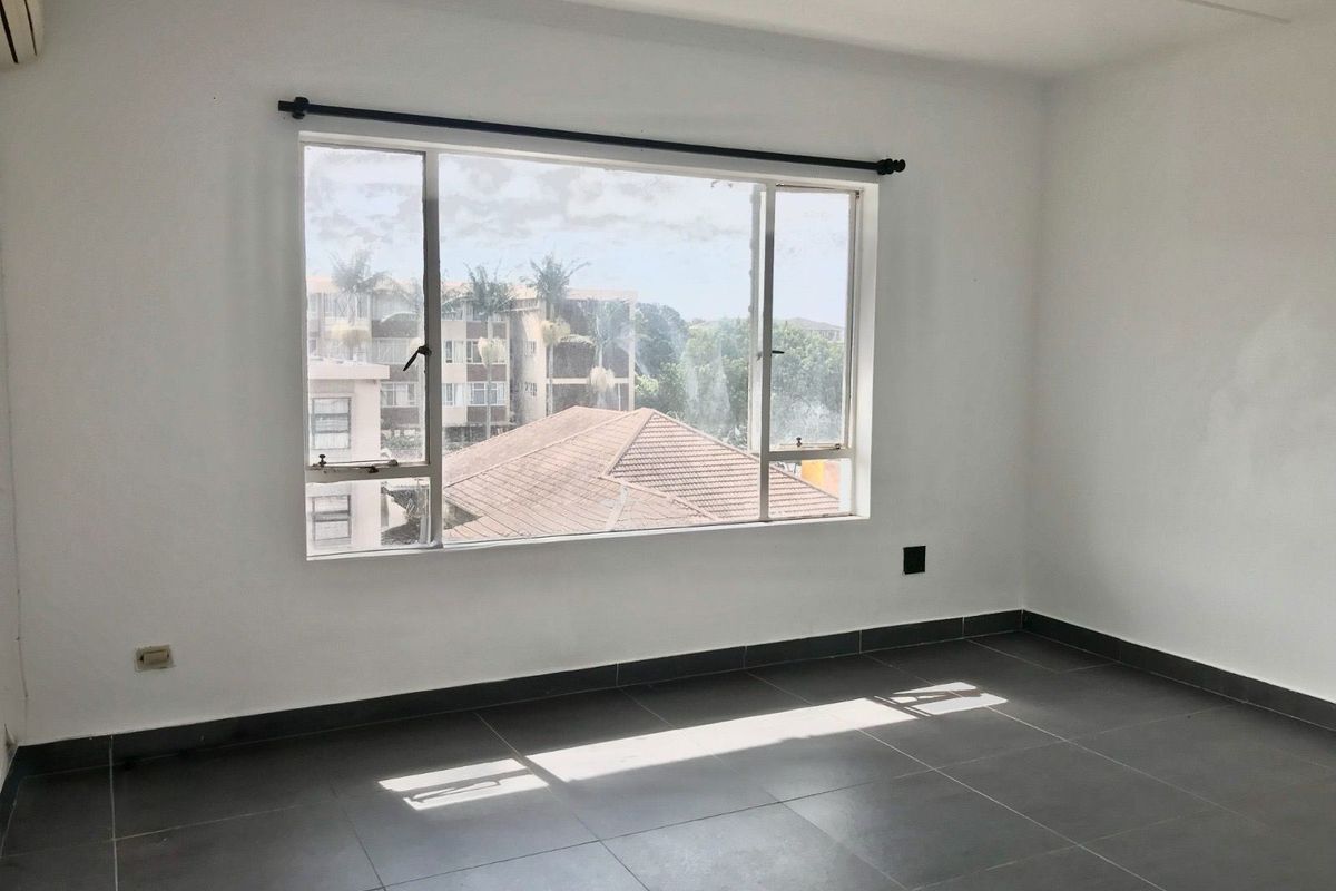 A bright, empty bedroom featuring large windows, neutral tiled flooring, and clean white walls, offering a versatile space ready for personalization.