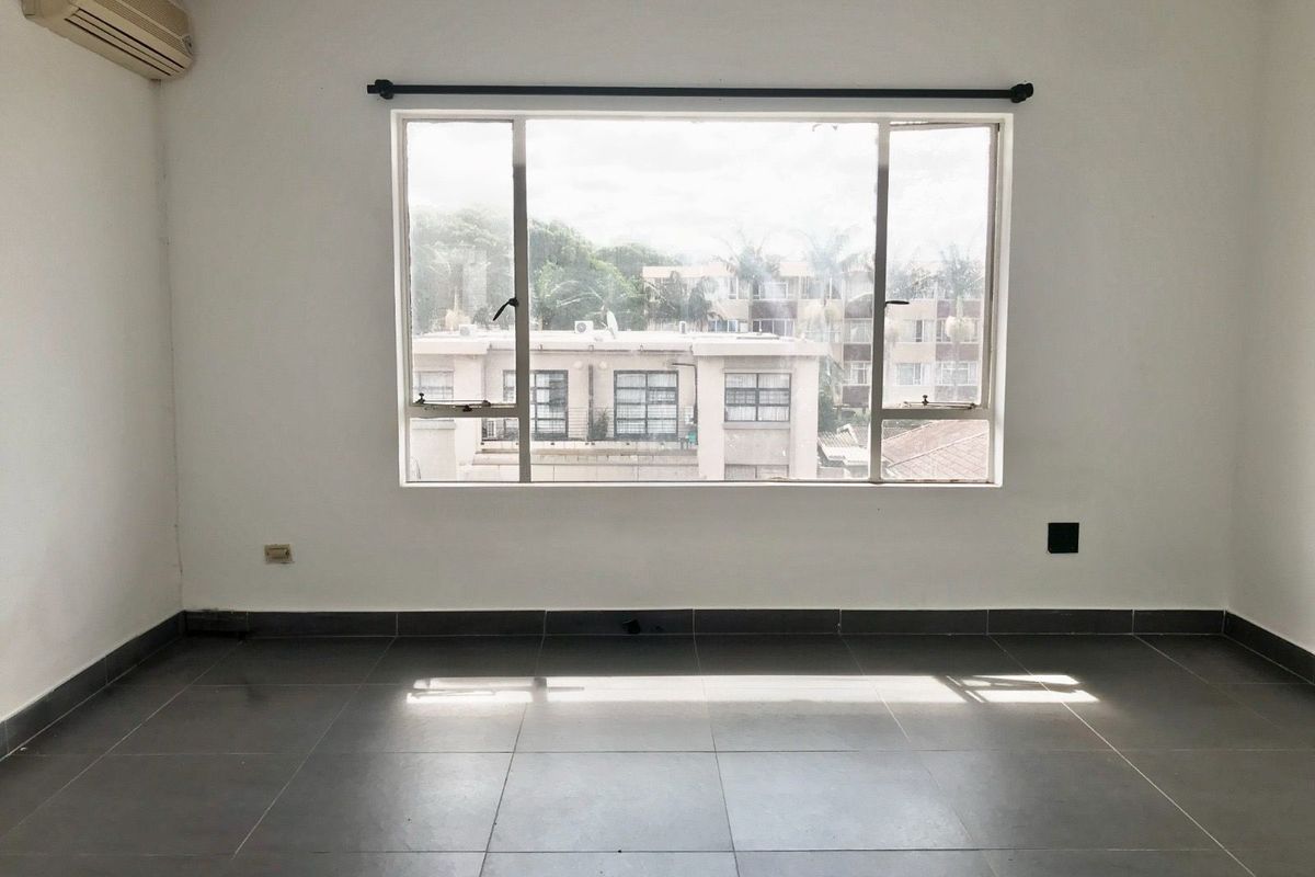 An empty, sunlit living room featuring large windows, dark floor tiles, and a wall-mounted air conditioning unit.