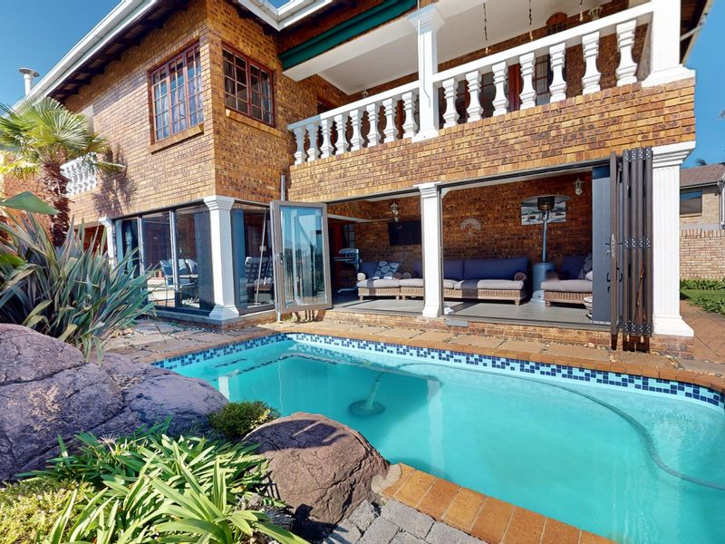 A well-maintained outdoor pool area featuring a brick facade, covered patio with folding glass doors, and a balcony with classic balustrades.