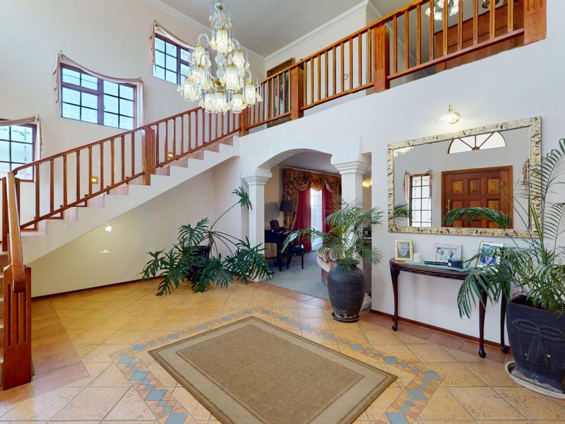 Spacious double-volume entrance hall featuring a grand wooden staircase, decorative columns, tiled flooring, and an elegant chandelier, leading into the main living areas.