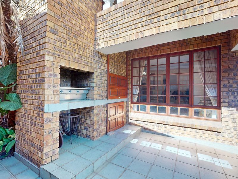 Covered patio area featuring a built-in brick braai, tiled flooring, and large timber-framed windows looking into the home.