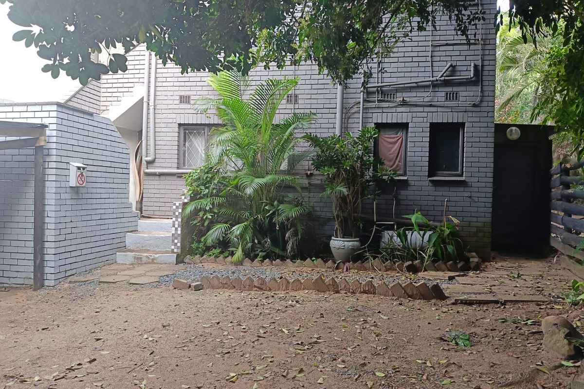 Rear exterior view of a grey brick house featuring external plumbing, concrete steps, and a small garden area.