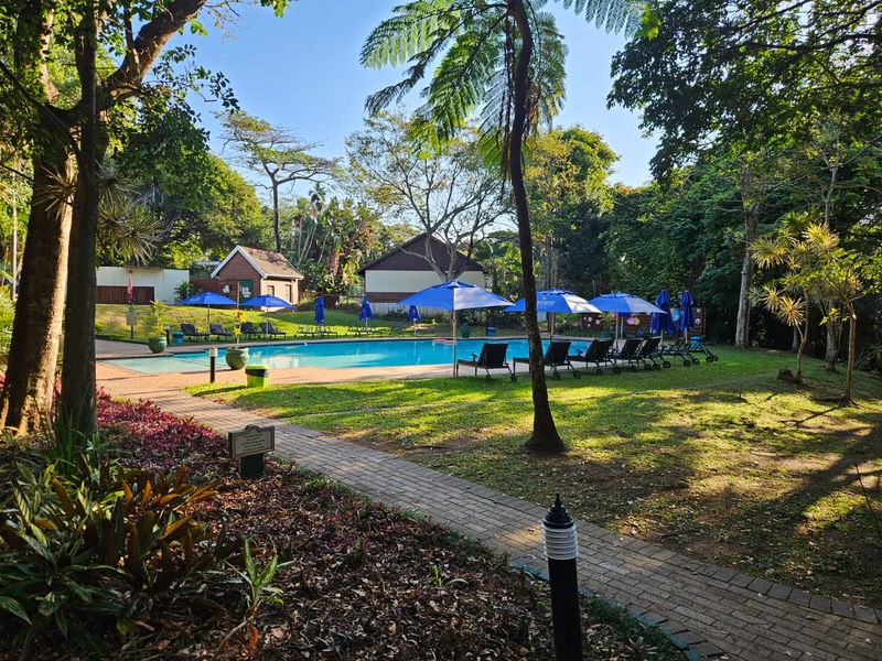 A well-maintained communal swimming pool area surrounded by lush landscaped gardens, mature trees, and paved walkways within the townhouse complex.
