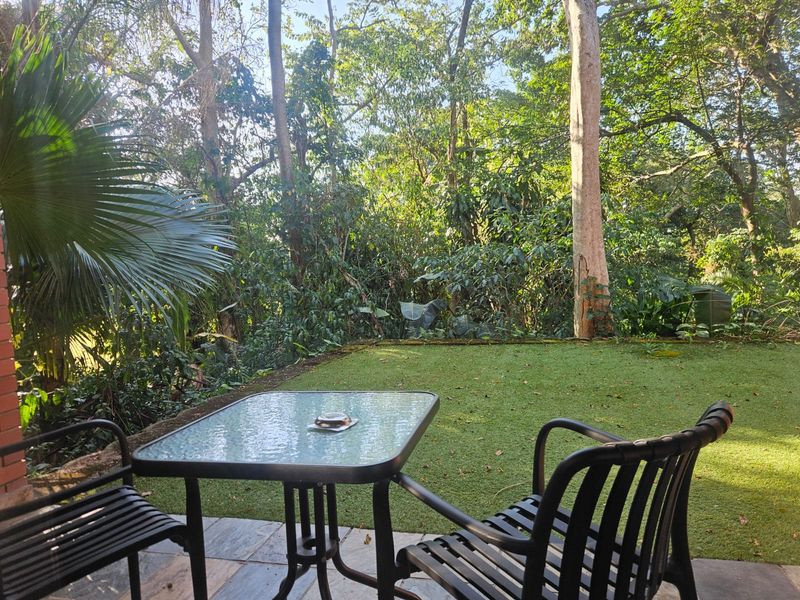 A private patio area featuring stone tile flooring and an adjacent artificial turf lawn, overlooking a lush, wooded garden setting.