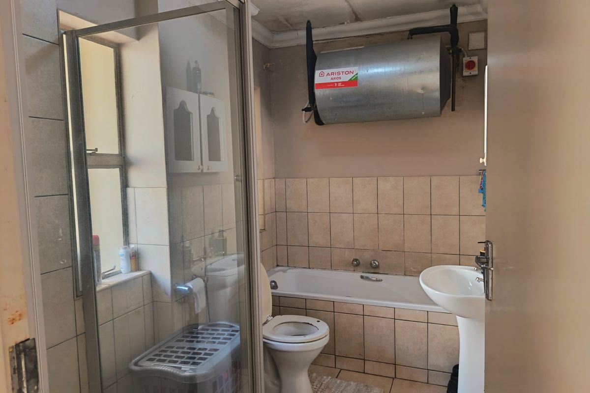 A functional bathroom featuring a shower enclosure, bathtub, pedestal basin, and a wall-mounted electric geyser, requiring cosmetic updates to improve its overall condition.