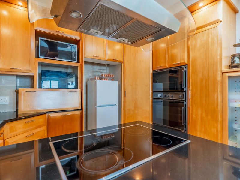 A functional kitchen featuring warm-toned wooden cabinetry, black granite countertops, a central electric hob, and integrated stainless steel appliances.