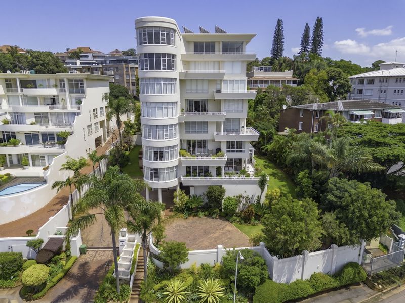 Aerial view of a multi-story penthouse building featuring a distinctive curved facade, private balconies, landscaped grounds, and a paved driveway entrance.