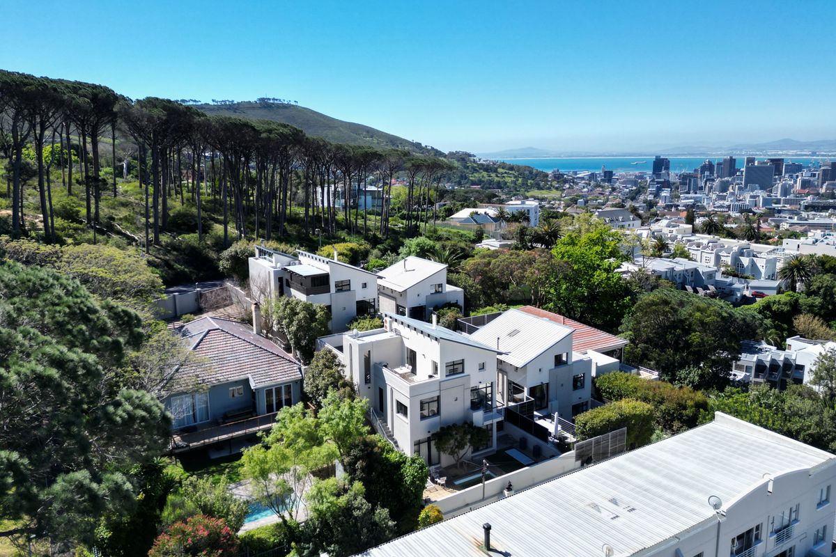 Aerial view of a modern residential property complex nestled against a forested hillside, featuring contemporary architecture, swimming pools, and panoramic city and ocean views.