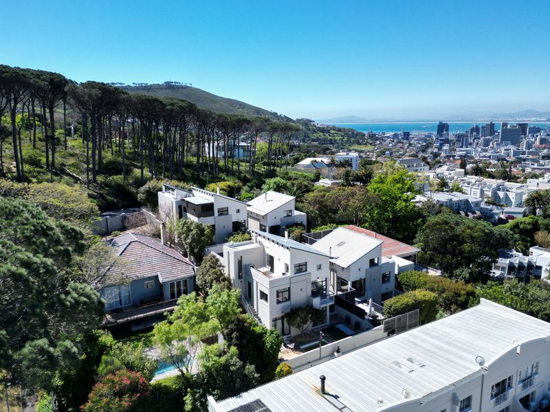 Aerial view of a modern residential property complex nestled against a forested hillside, featuring contemporary architecture, swimming pools, and panoramic city and ocean views.