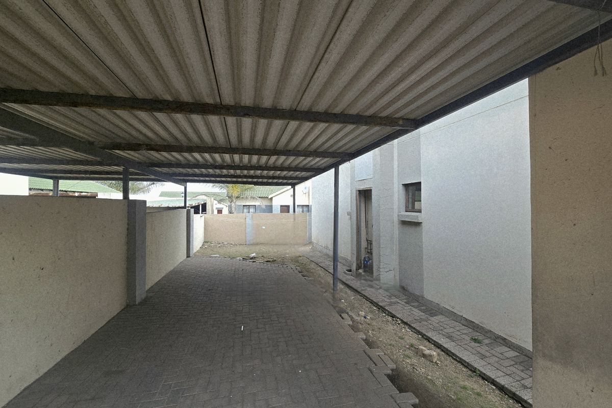 A covered carport area featuring a corrugated metal roof, paved flooring, and surrounding boundary walls, providing sheltered off-street parking.