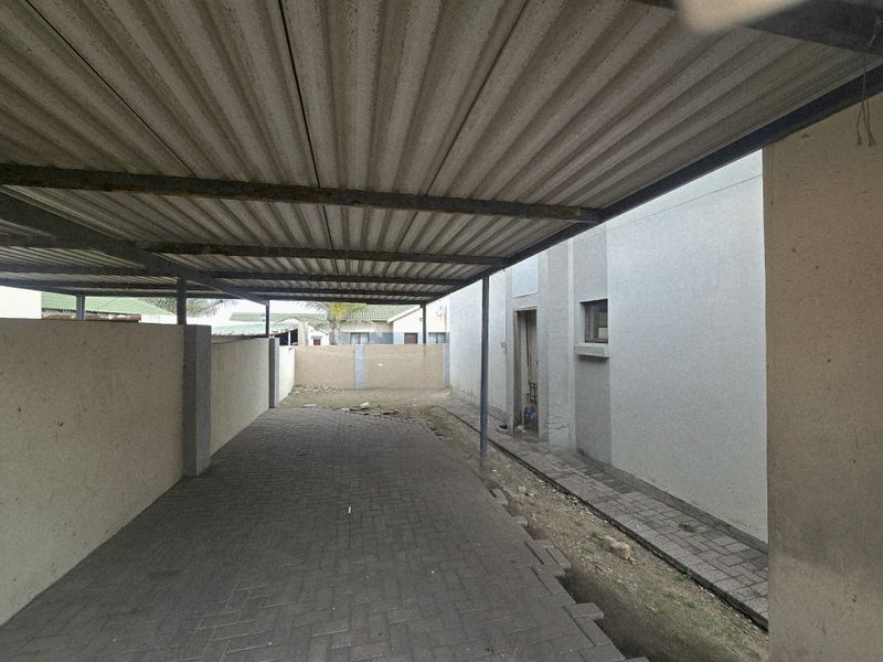 A covered carport area featuring a corrugated metal roof, paved flooring, and surrounding boundary walls, providing sheltered off-street parking.