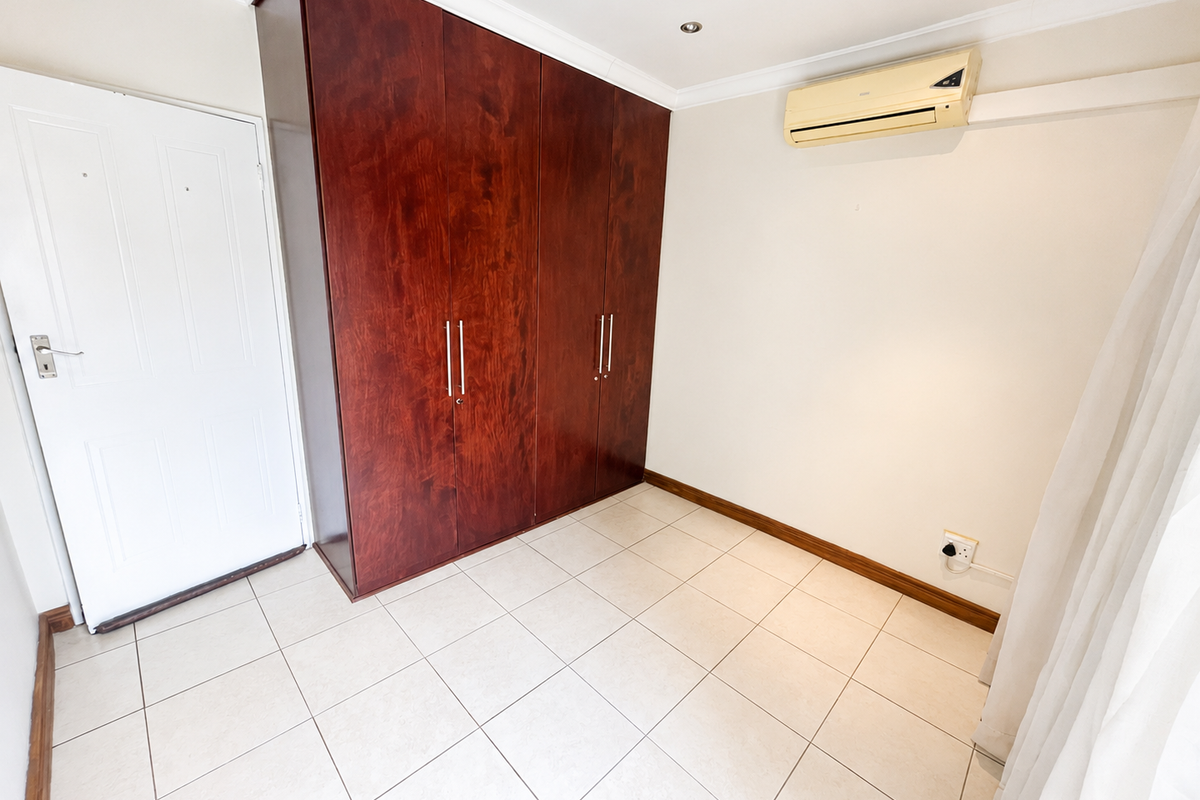 A clean, empty bedroom featuring tiled flooring, a large built-in wooden wardrobe, and a wall-mounted air conditioning unit.