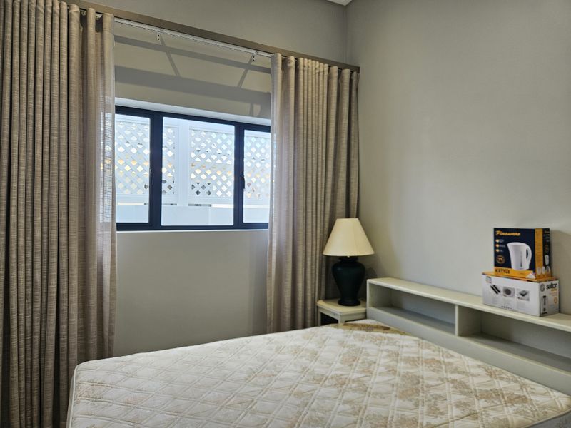 A well-maintained bedroom featuring a built-in headboard with integrated shelving, neutral wall tones, and large windows with floor-to-ceiling curtains.