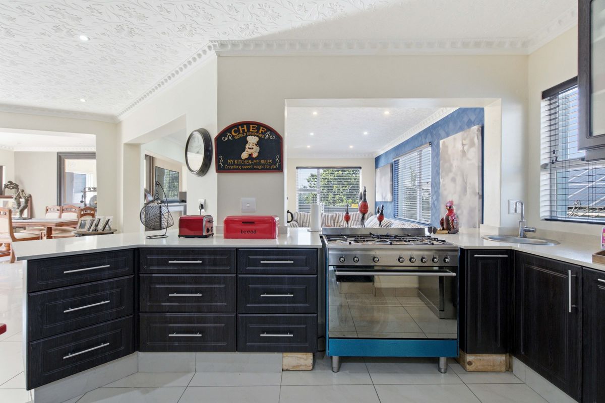 A functional kitchen featuring dark cabinetry, stone countertops, a gas stove with oven, and tiled flooring, opening into an adjacent living area.