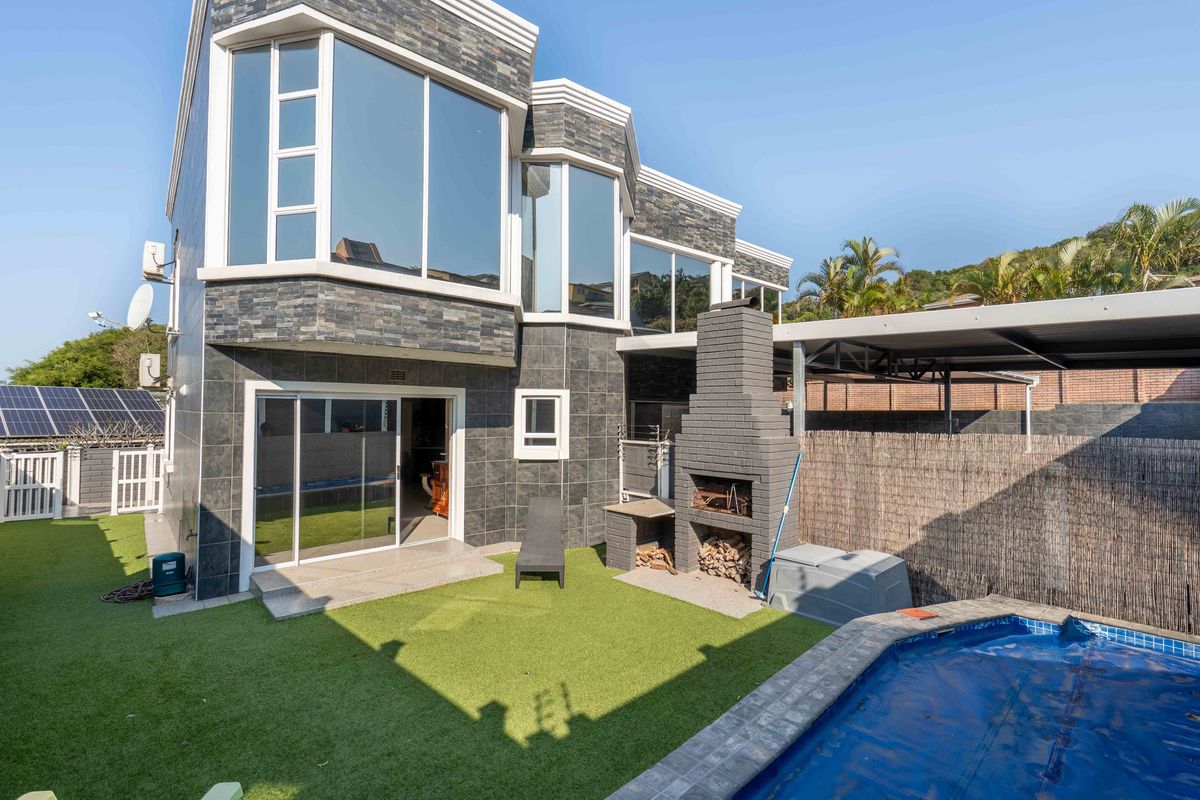 Rear exterior view featuring stone-clad facade, built-in braai area, swimming pool with cover, and artificial lawn in a well-maintained garden setting.