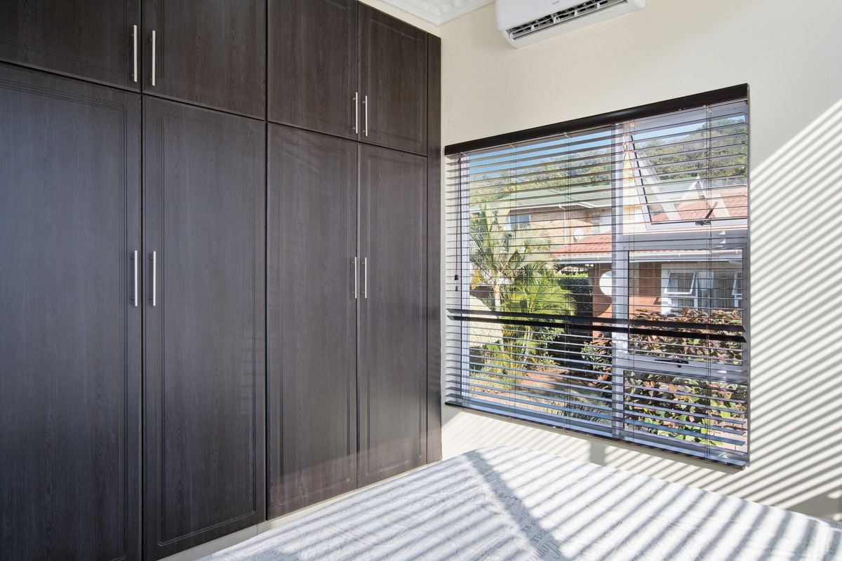 A bright bedroom featuring dark wood built-in cupboards, an air conditioning unit, and venetian blinds, offering a clean and functional space.