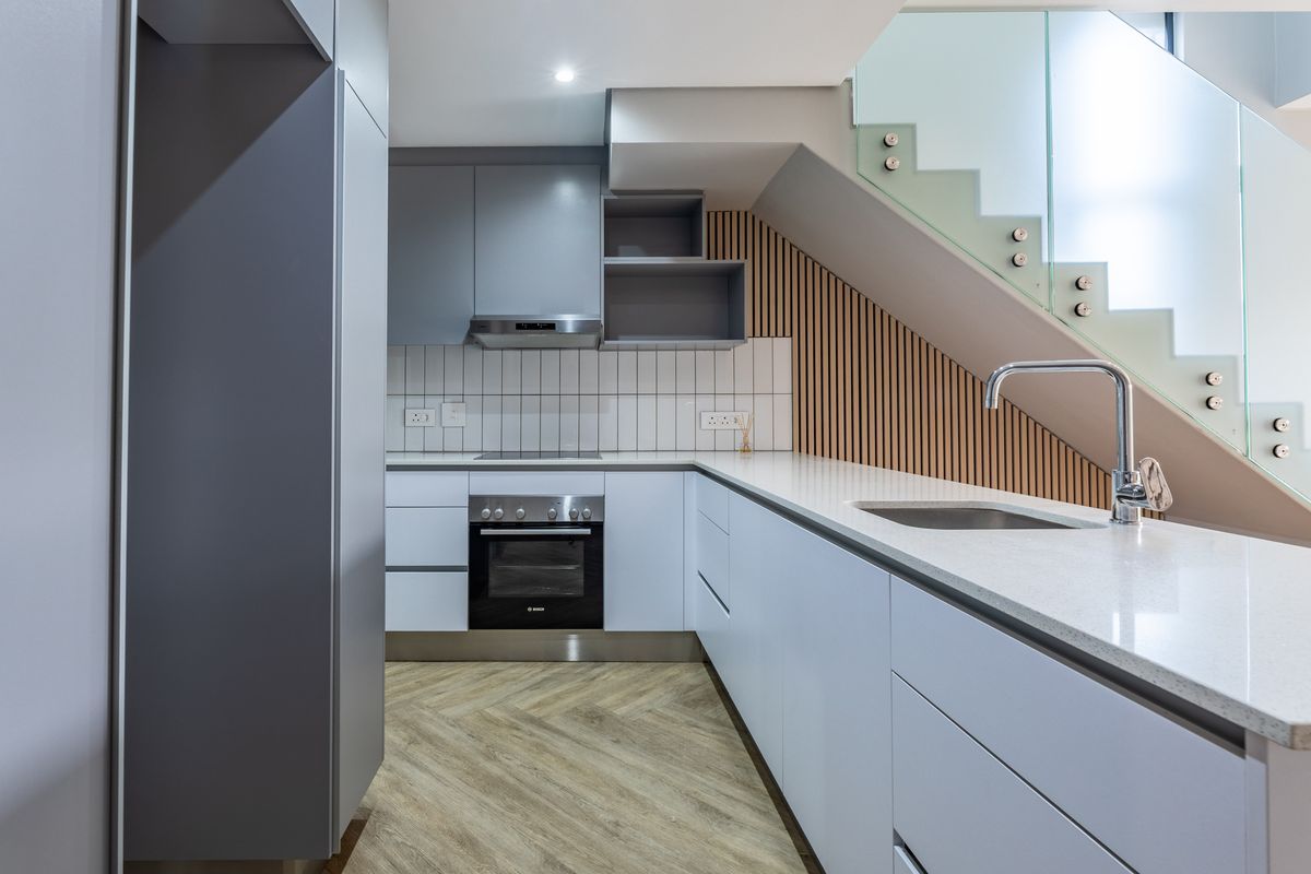 Modern kitchen featuring sleek grey cabinetry, quartz countertops, integrated appliances, and a stylish herringbone floor, positioned beneath a contemporary glass-railed staircase.