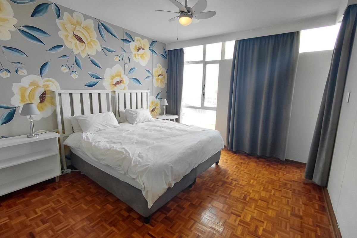 A well-lit bedroom featuring classic parquet flooring, a ceiling fan, and large windows with floor-to-ceiling curtains.