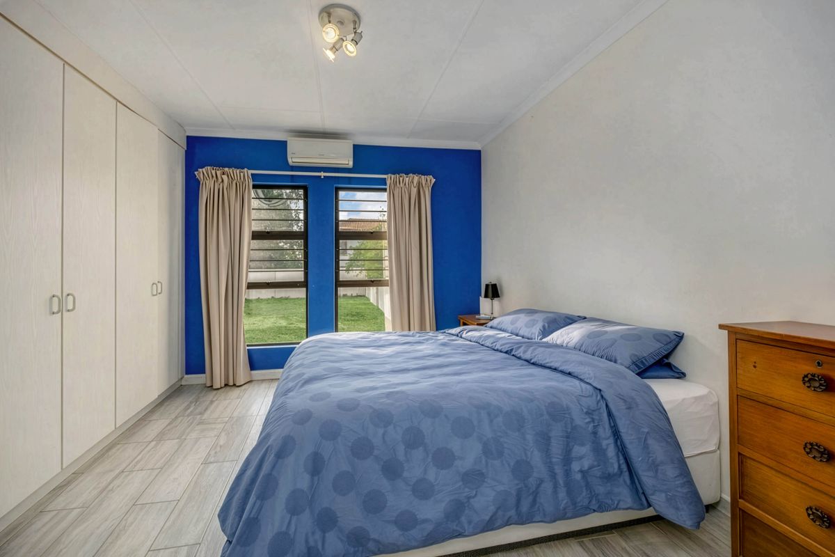 A well-lit bedroom featuring built-in wardrobes, tiled flooring, a wall-mounted air conditioning unit, and large windows overlooking the garden.