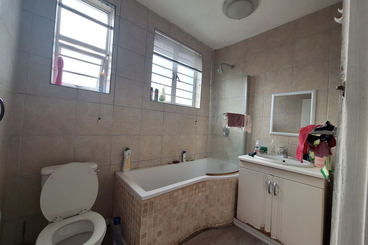 A functional bathroom featuring a bathtub with shower screen, vanity basin, and toilet, finished with neutral wall tiling and louver windows.