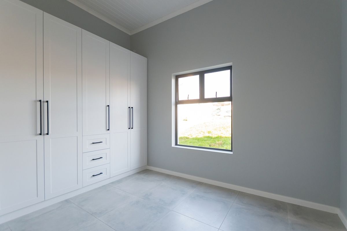 A bright, modern bedroom featuring neutral grey walls, large format floor tiles, and extensive white built-in cabinetry with contemporary black hardware.