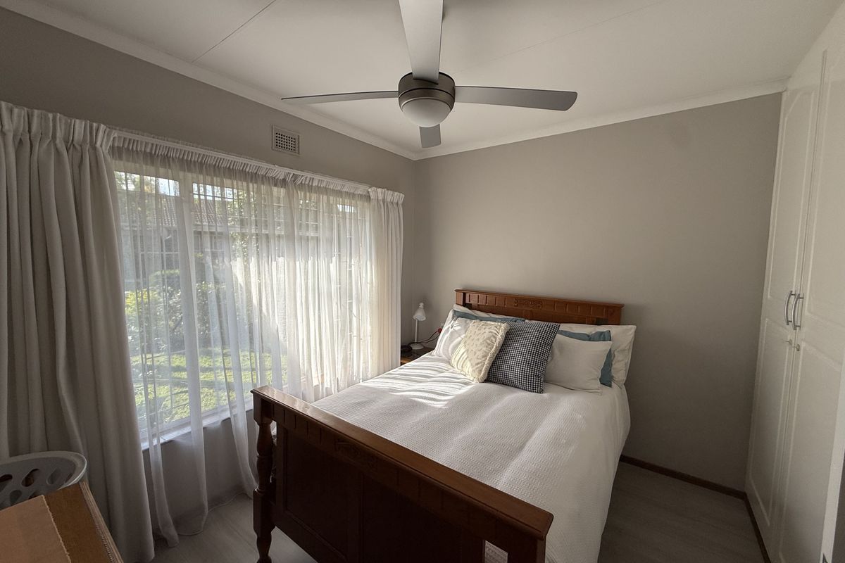 A well-maintained bedroom featuring built-in wardrobes, a ceiling fan, and large windows with sheer curtains allowing for natural light.