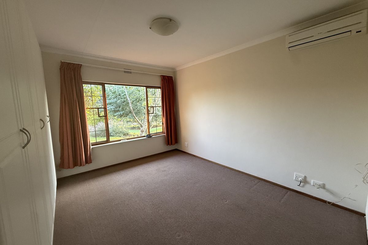 A spacious, empty bedroom featuring built-in cupboards, carpeted flooring, and a wall-mounted air conditioning unit with a view of the garden.
