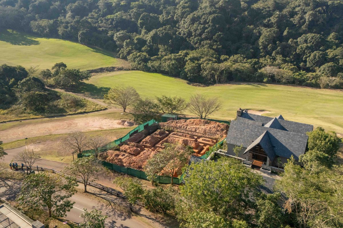 Aerial view of a residential property featuring a pitched roof house adjacent to an active construction site and expansive green landscape.